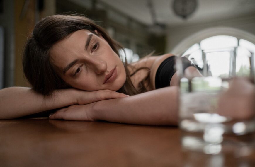 Pensive woman resting her head on a wooden table with a glass of water nearby, indoors.