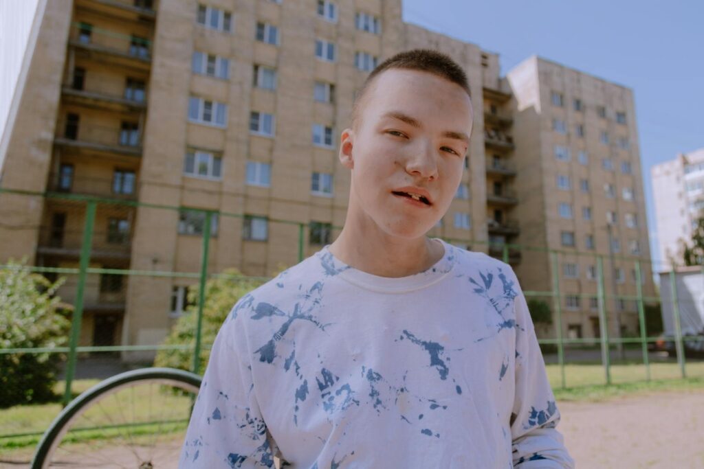 Young male teenager stands outdoors with a casual expression and urban backdrop.