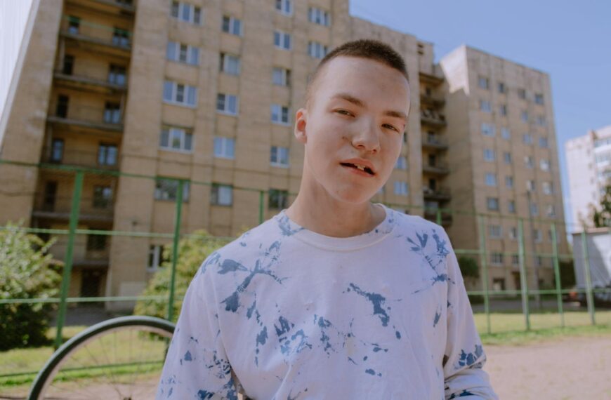 Young male teenager stands outdoors with a casual expression and urban backdrop.