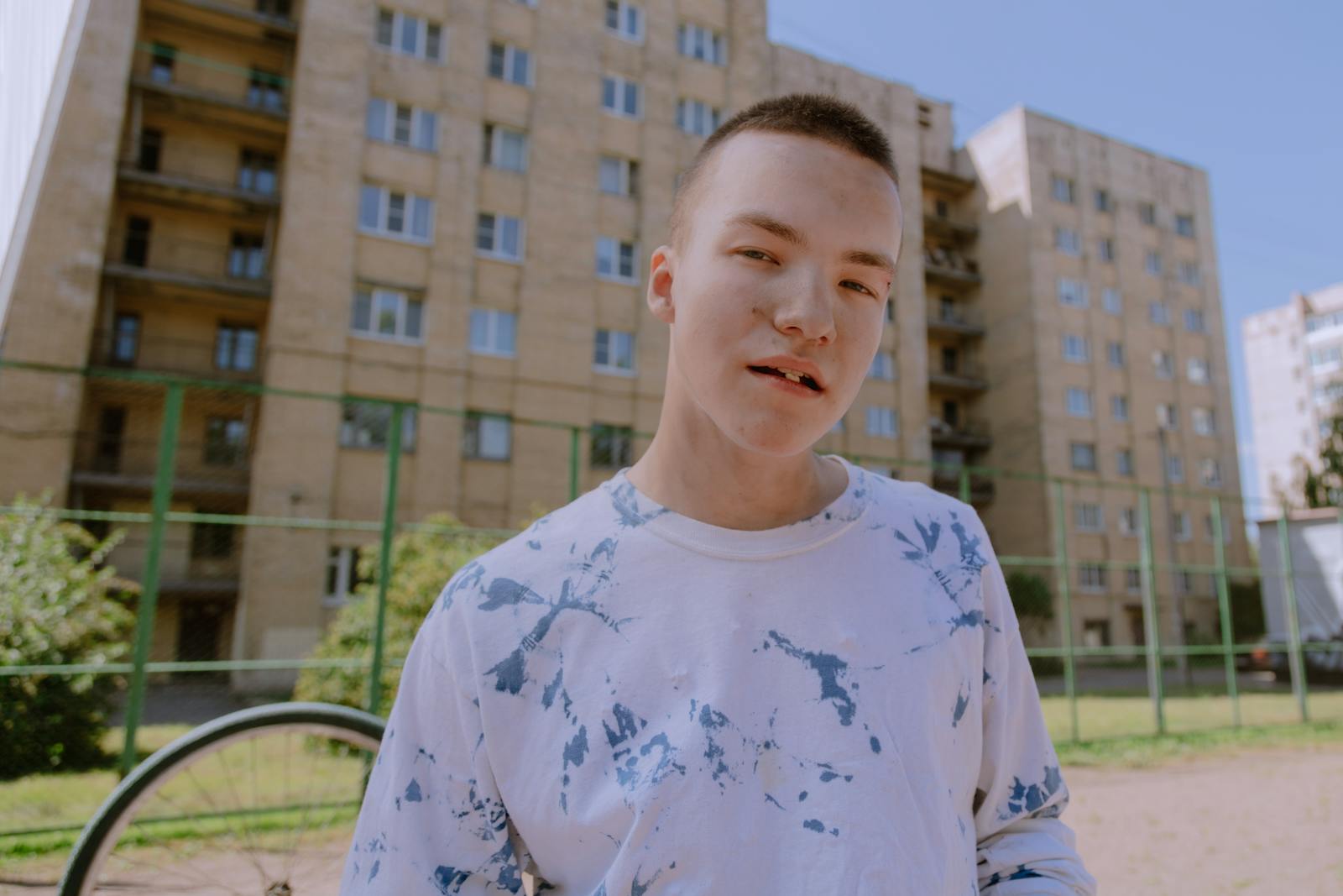 Young male teenager stands outdoors with a casual expression and urban backdrop.