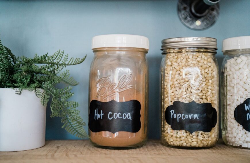Glass jars with popcorn, hot cocoa, and marshmallows on a kitchen shelf with a potted plant.
