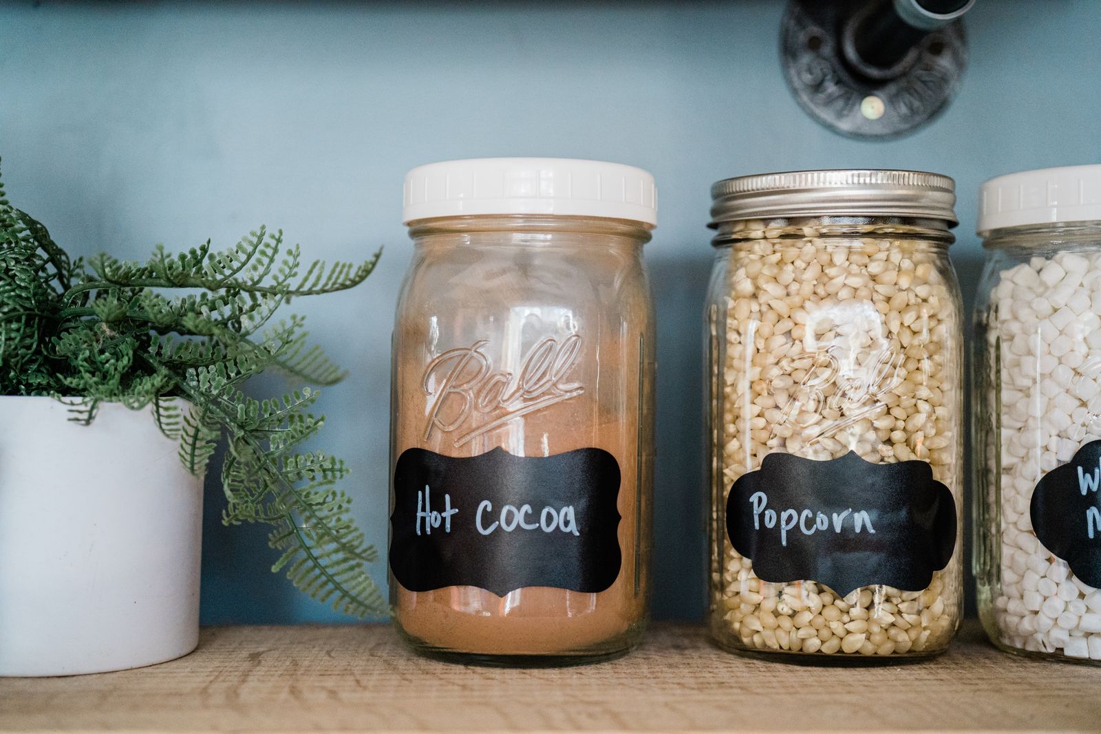 Glass jars with popcorn, hot cocoa, and marshmallows on a kitchen shelf with a potted plant.