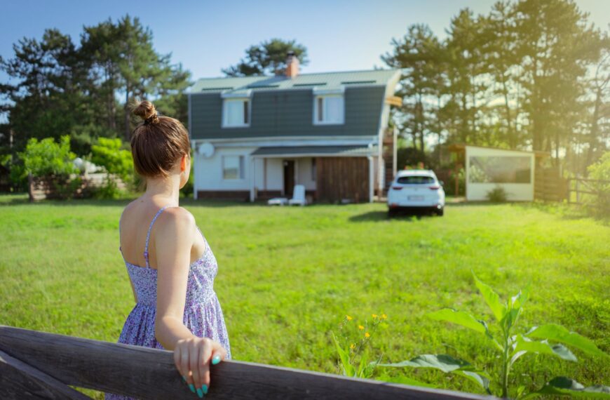 A woman in a sundress stands by a fence admiring a countryside house on a sunny day.