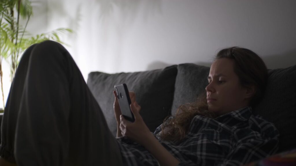A woman sits on a sofa, focused on her smartphone in a dimly lit room, creating a relaxed ambiance.