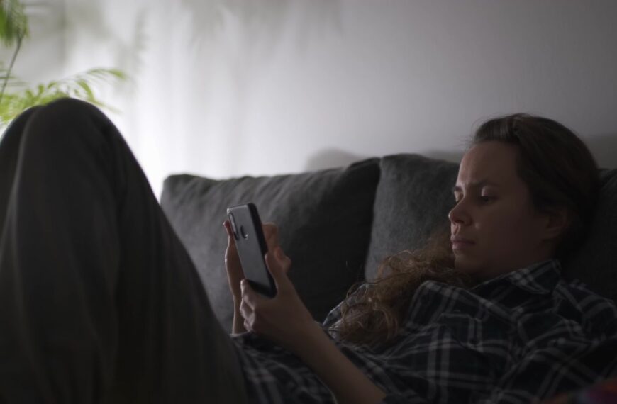 A woman sits on a sofa, focused on her smartphone in a dimly lit room, creating a relaxed ambiance.