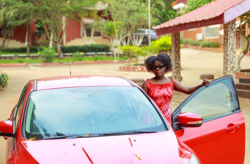 A fashionable woman in red attire stands with a red car in an outdoor setting.