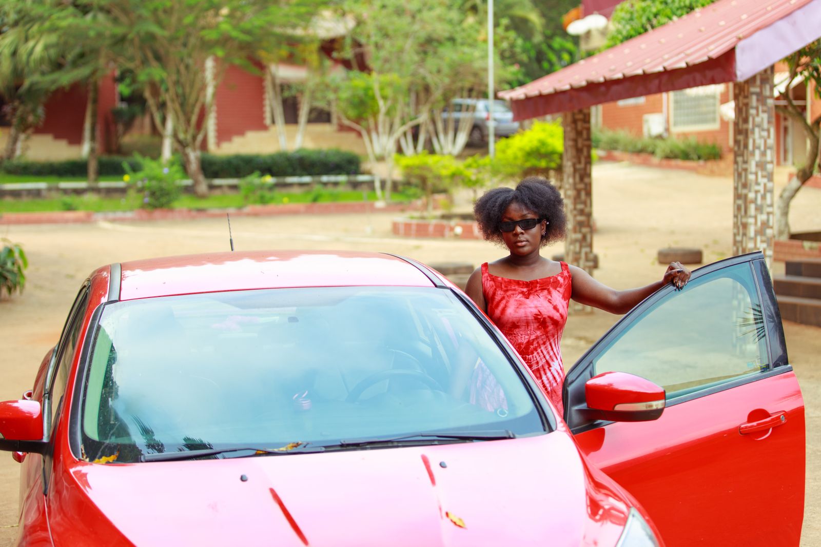 A fashionable woman in red attire stands with a red car in an outdoor setting.