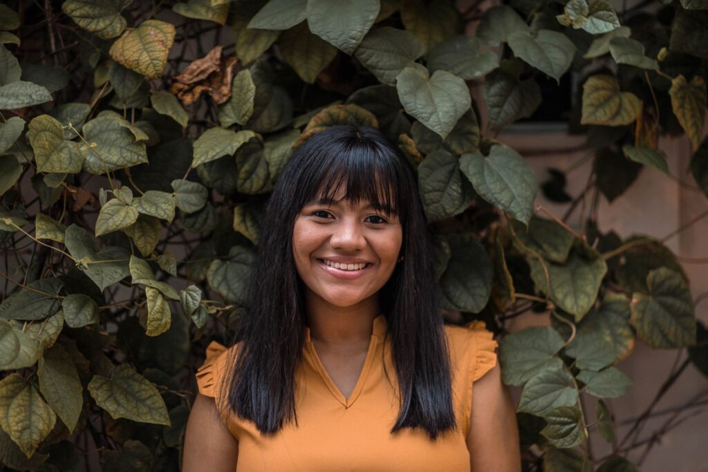 A cheerful woman stands against a backdrop of lush green leaves, radiating positivity.