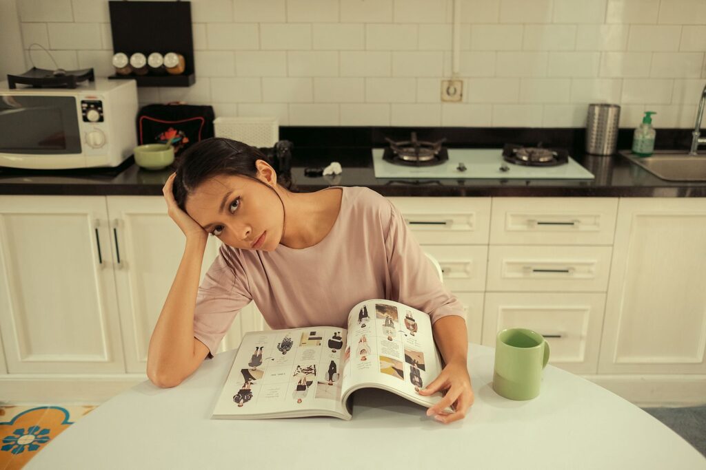 Woman sitting in a kitchen, reading a magazine at a table with a thoughtful expression.