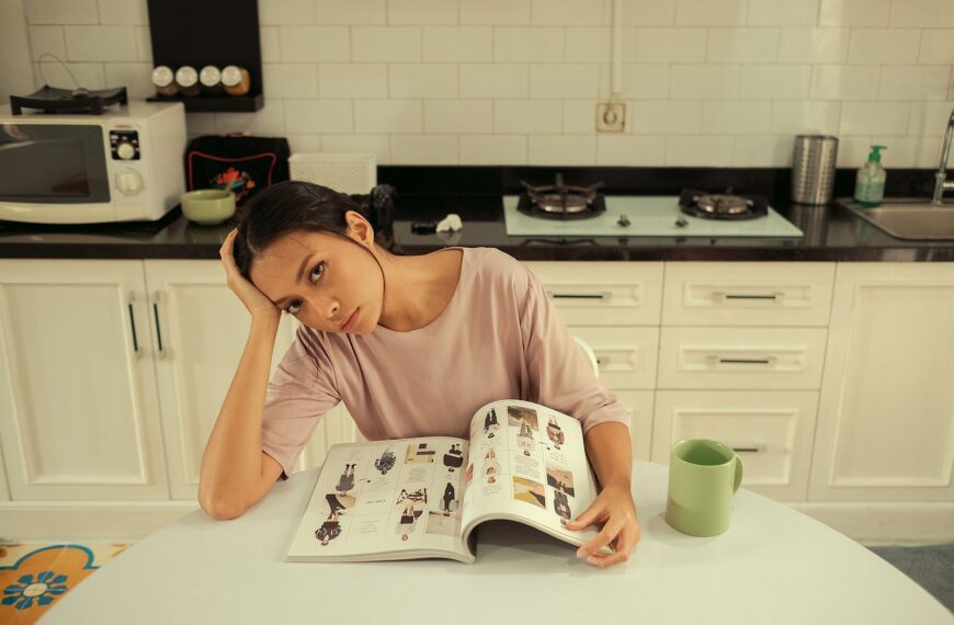 Woman sitting in a kitchen, reading a magazine at a table with a thoughtful expression.