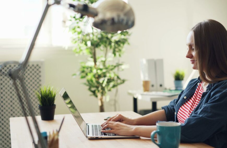 Woman smiling while working on a laptop in a modern office with plants.