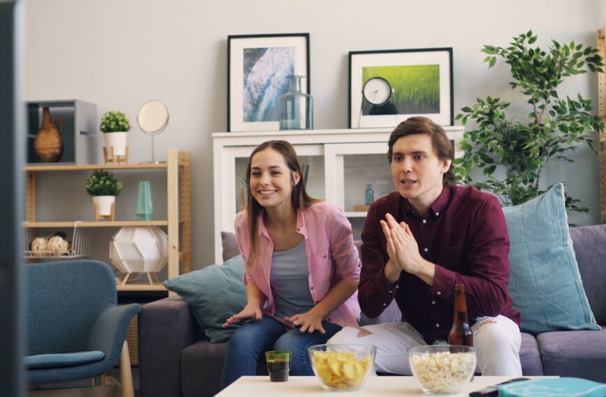 A young couple excitedly watching TV at home, enjoying snacks together in a lively living room setting.