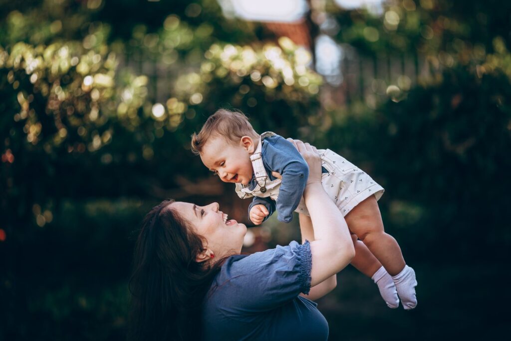 A joyful mother lifts her smiling baby outdoors in a summer park, capturing a loving moment.