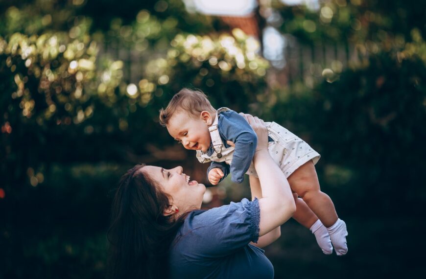 A joyful mother lifts her smiling baby outdoors in a summer park, capturing a loving moment.