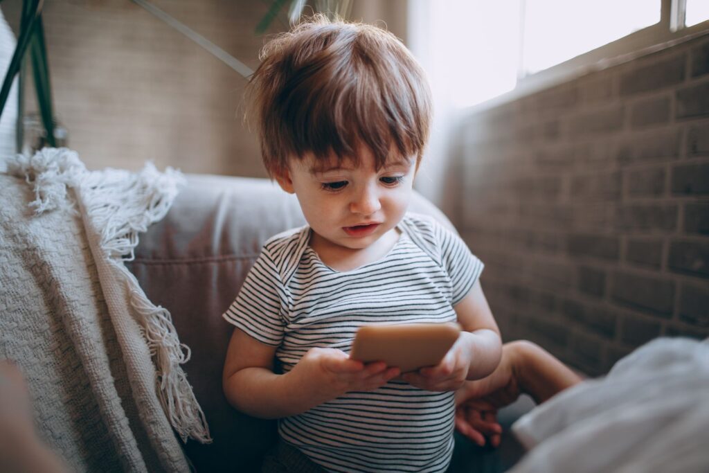 A cute toddler in a striped shirt focuses on a smartphone indoors, near a window.