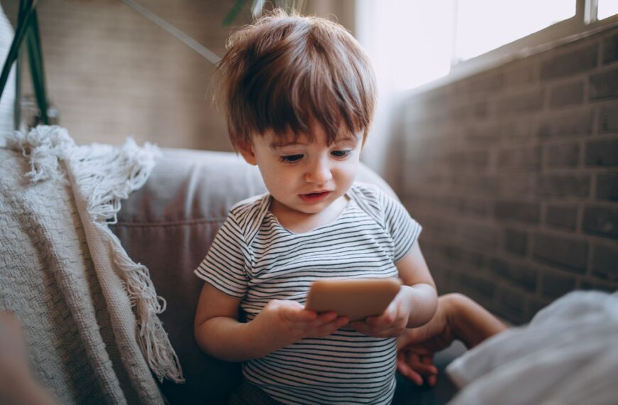 A cute toddler in a striped shirt focuses on a smartphone indoors, near a window.