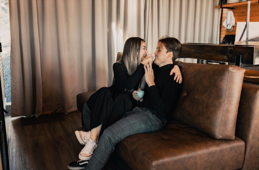A happy couple laughing and enjoying coffee together on a cozy indoor sofa setting.