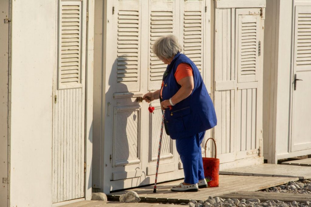 Senior woman with cane opens beach hut door on sunny day.