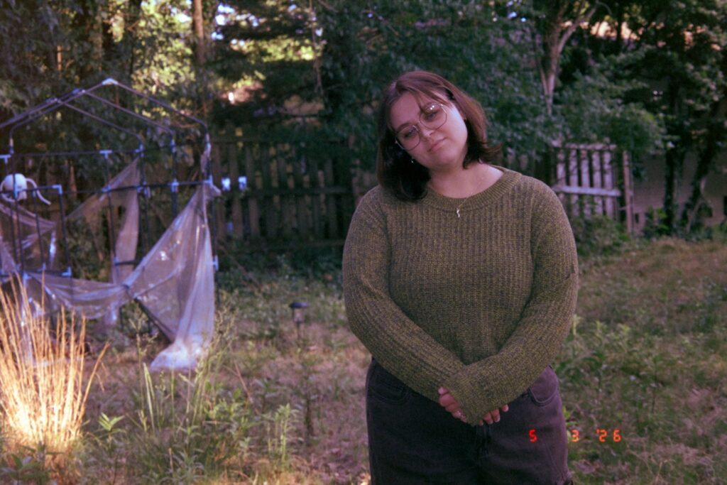 Woman in green sweater stands in a rustic outdoor setting.