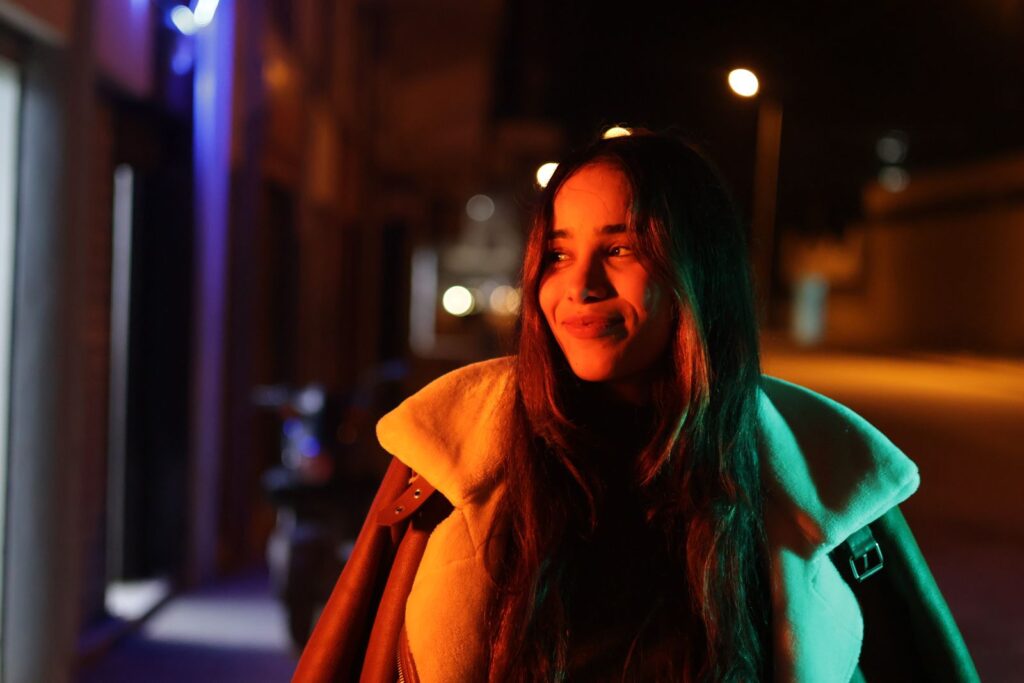 Smiling woman in a coat captured on a city street at night with vibrant lighting.