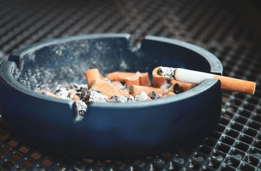 Close-up of cigarette butts and ashes in a blue ashtray with a smoldering cigarette, highlighting smoking habits.