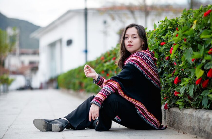 A young woman sits on pavement in a colorful poncho, outdoors beside bushes.