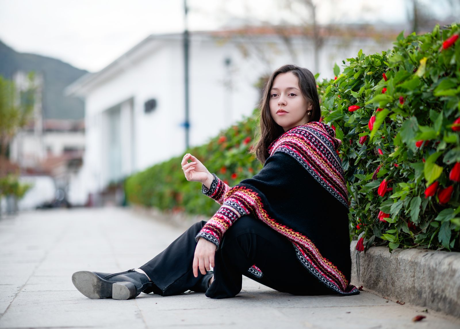 A young woman sits on pavement in a colorful poncho, outdoors beside bushes.