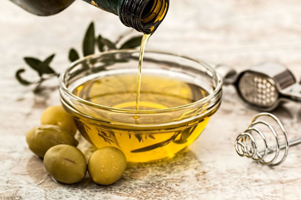 Close-up of olive oil being poured into a glass bowl surrounded by fresh olives and kitchen tools.
