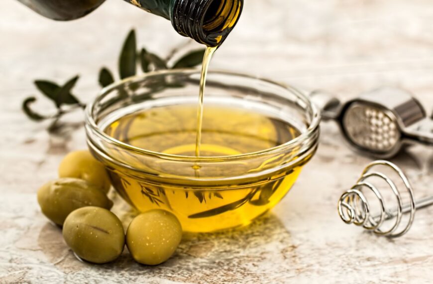 Close-up of olive oil being poured into a glass bowl surrounded by fresh olives and kitchen tools.