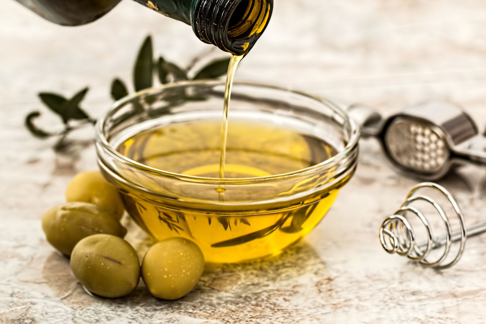 Close-up of olive oil being poured into a glass bowl surrounded by fresh olives and kitchen tools.