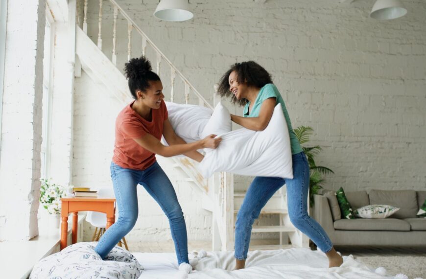 Two women having a playful pillow fight in a sunlit modern living room.