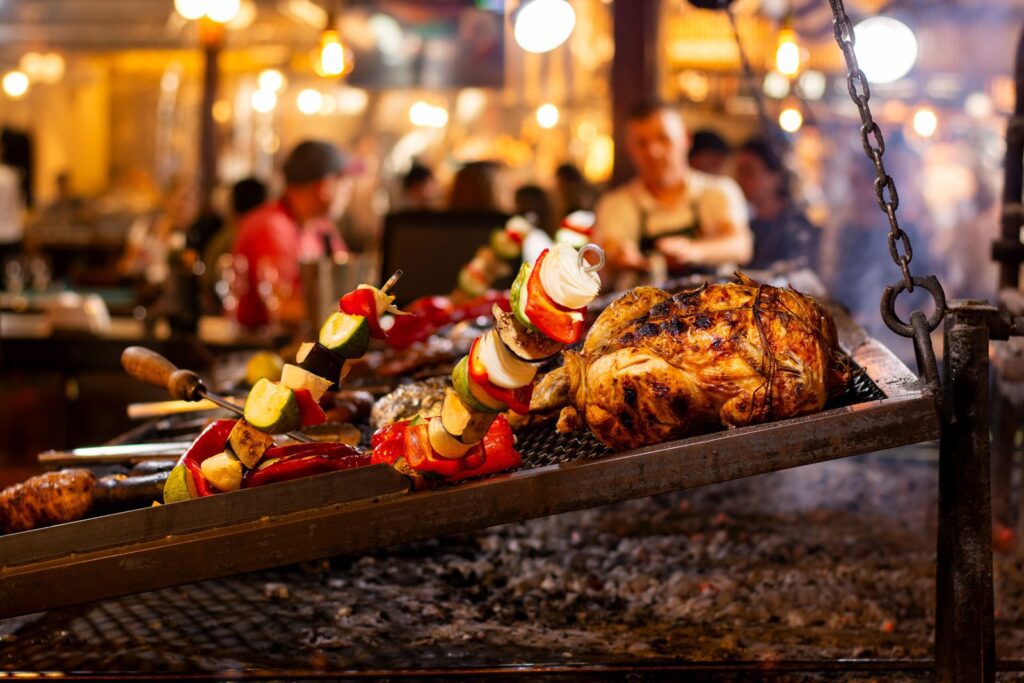 Grilled chicken and vegetable skewers over an open flame in a lively market setting.