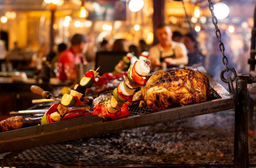 Grilled chicken and vegetable skewers over an open flame in a lively market setting.