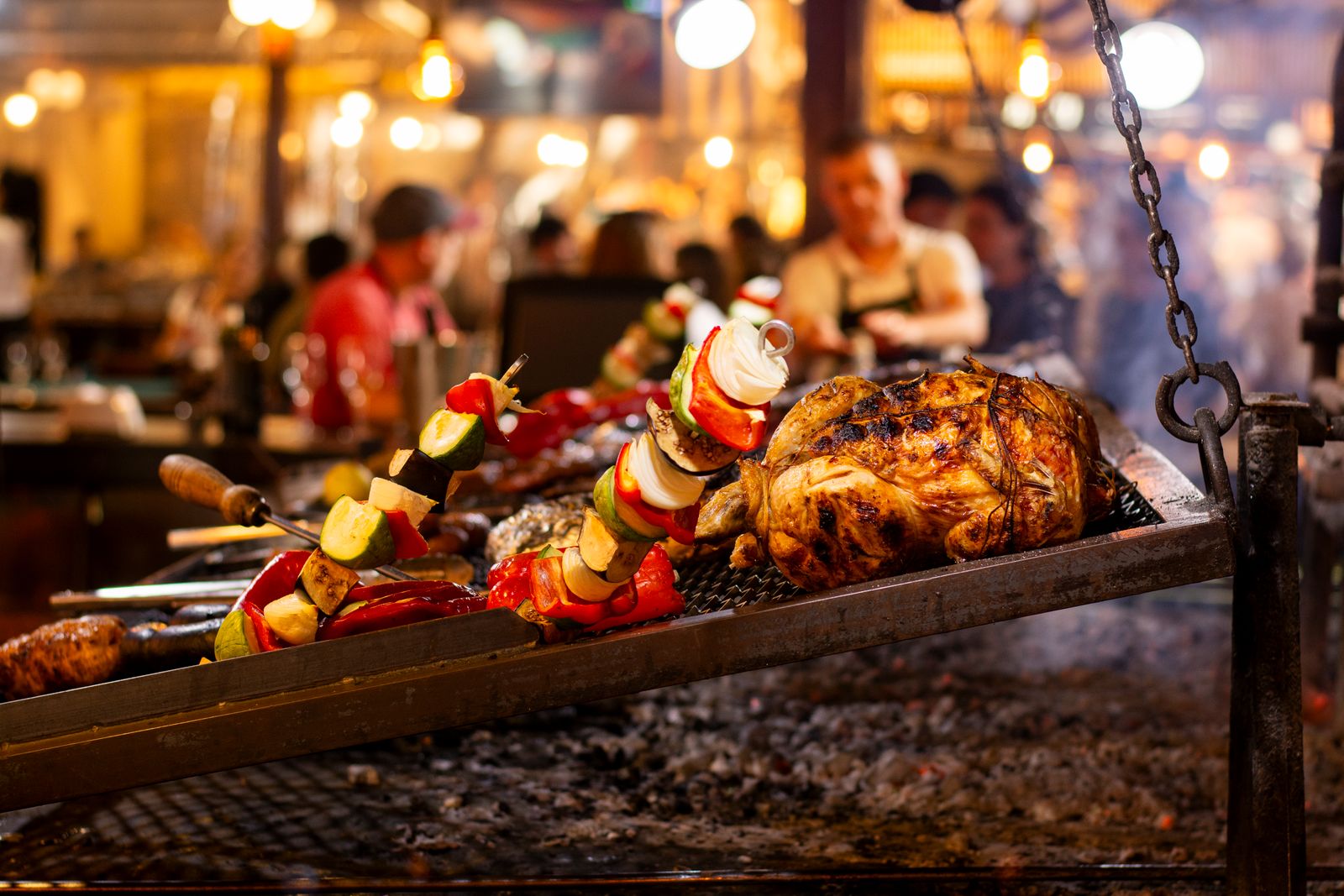 Grilled chicken and vegetable skewers over an open flame in a lively market setting.