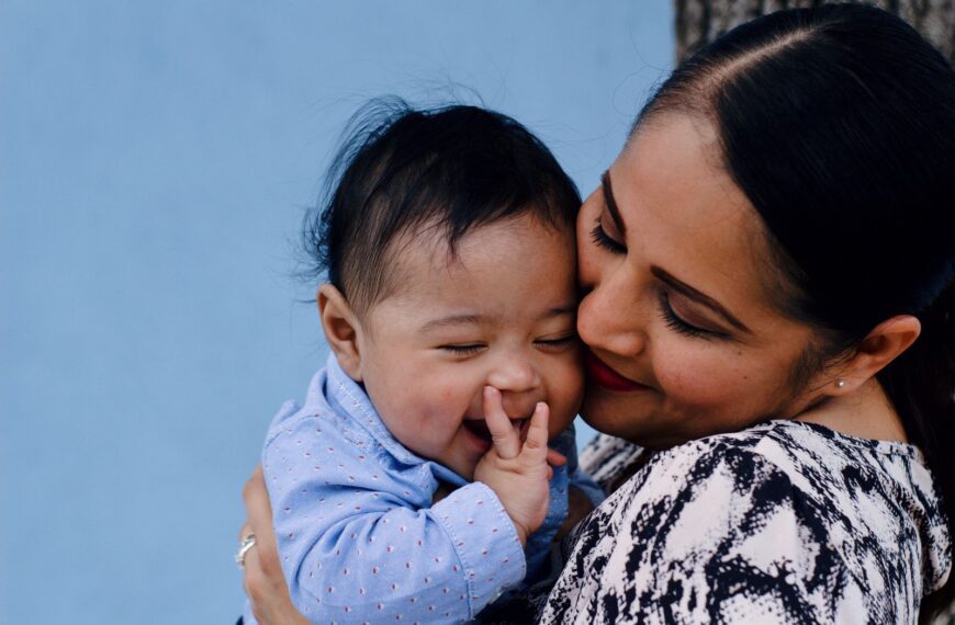 A mother affectionately embraces her baby against a serene blue background.