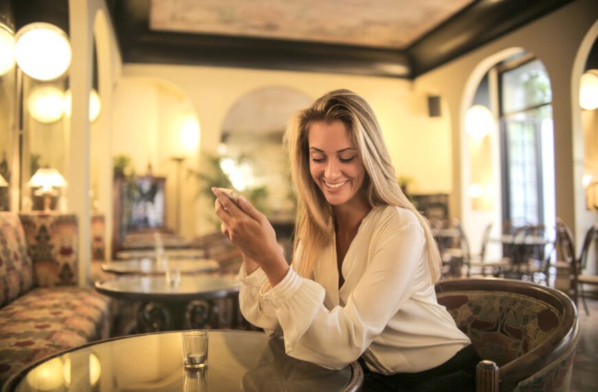 Smiling elegant woman in white blouse using mobile phone while sitting at table in old fashioned pub with glass shot on table