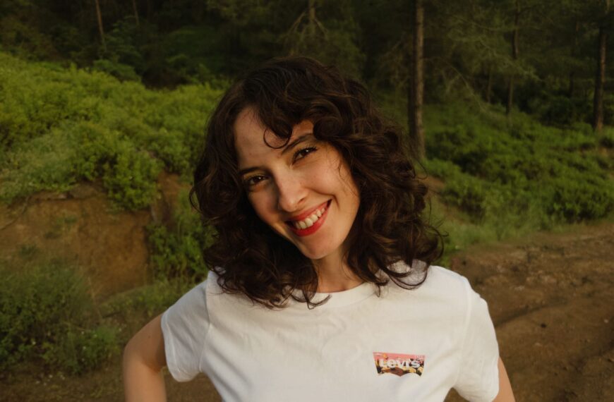 A cheerful woman with curly hair smiling outdoors in a green forest setting.