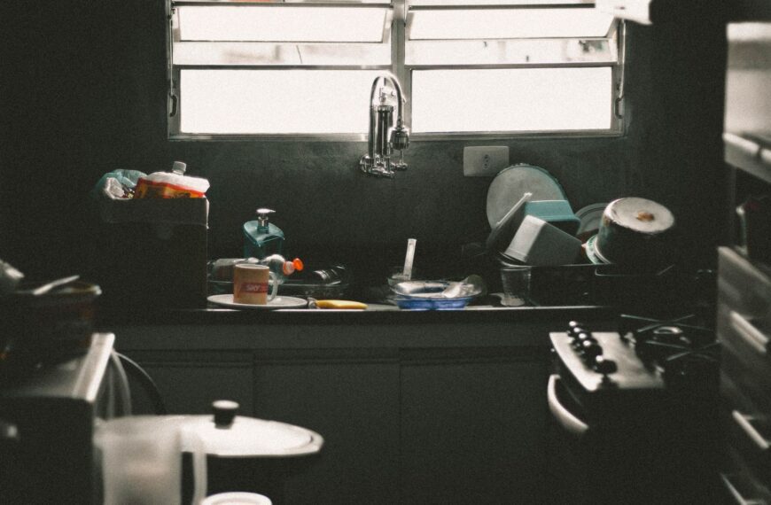 A cluttered kitchen sink filled with dirty dishes and utensils near a window, creating a messy home interior scene.