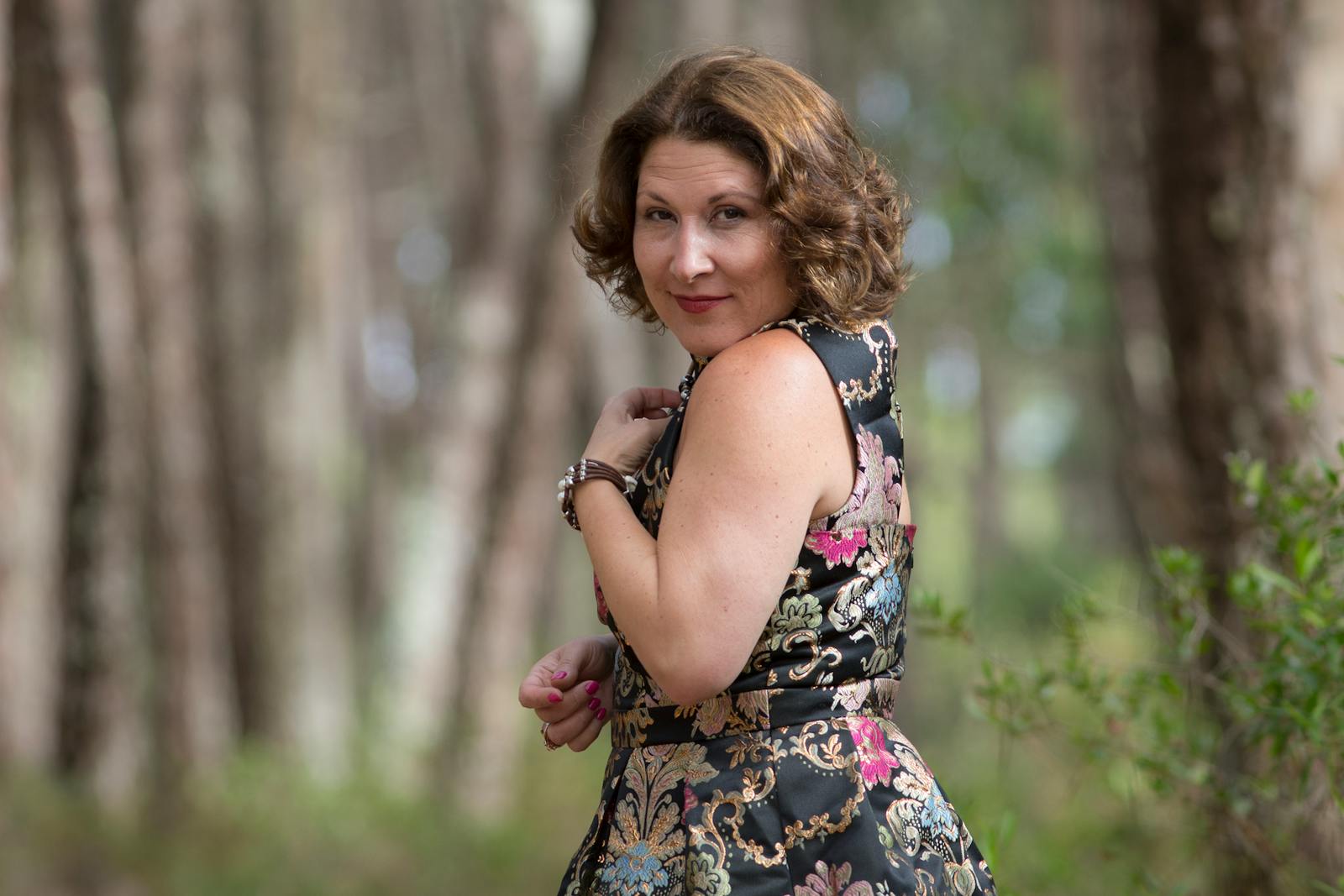 Woman in a stylish floral dress poses in a serene forest setting, capturing a moment of elegance.