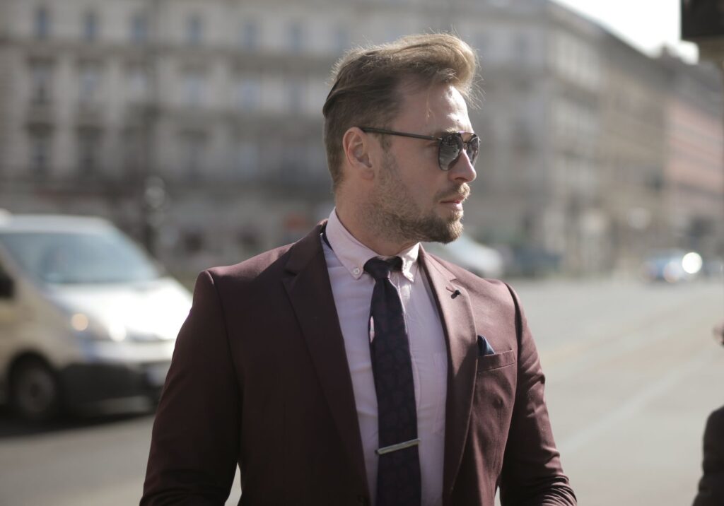 Businessman in sunglasses wearing maroon suit and tie walks confidently in urban setting.