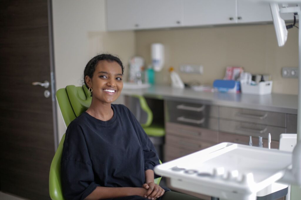Positive young African American female in casual wear sitting on green dental chair near table with medical tools while waiting for stomatologist and looking away in daylight