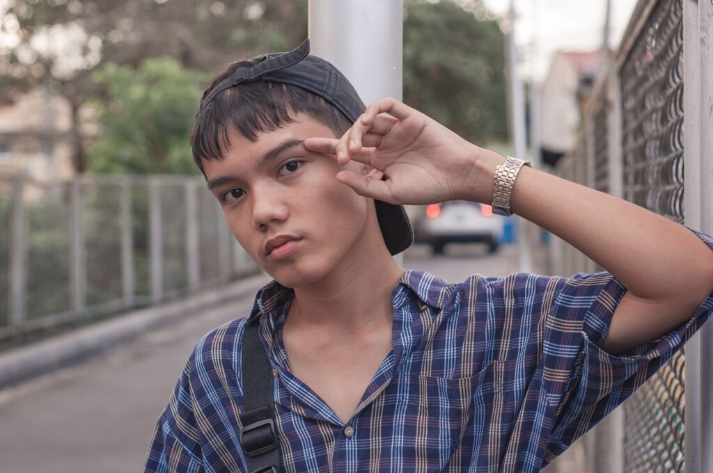 Ethnic male teenager in casual clothes and cap standing near metal mesh fence on street and looking at camera