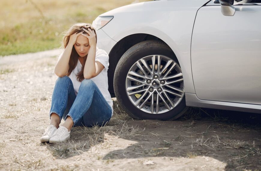 Frustrated female driver in white t shirt and jeans sitting on ground near damaged car with hands on head during car travel in sunny summer day