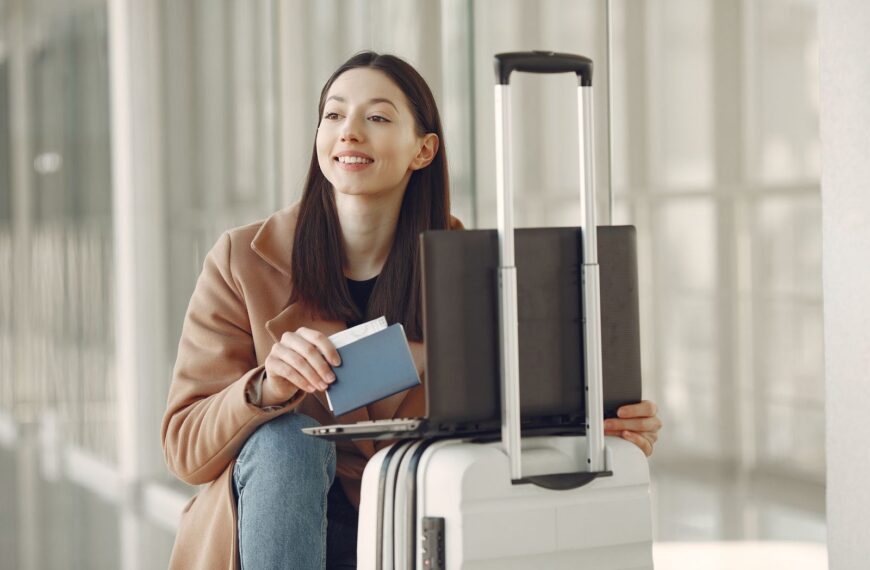 Smiling adult ethnic female traveler in trendy coat holding passport and using laptop on luggage in airport corridor for checking ticket for correctness