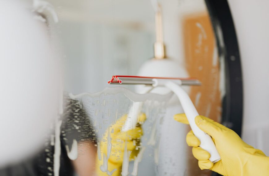 Anonymous person in yellow rubber gloves wiping water by squeegee from mirror in bathroom