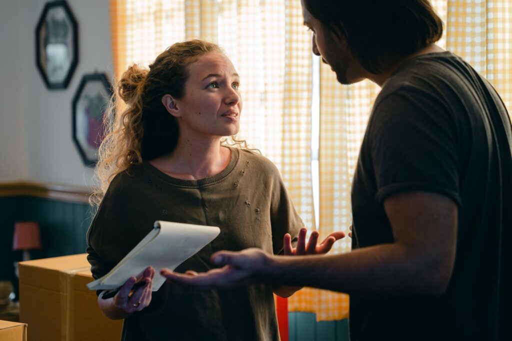 Young perplexed couple wearing casual clothes with notebook looking at each other nervously while unpacking carton box