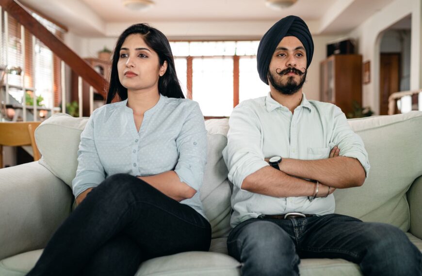 Resentful young Indian couple ignoring each other while sitting on couch together with crossed arms