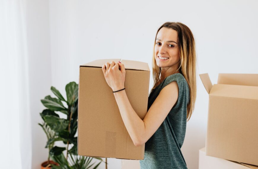 Side view smiling lady in casual green shirt carrying cardboard box while preparing for moving in new home and looking at camera happily