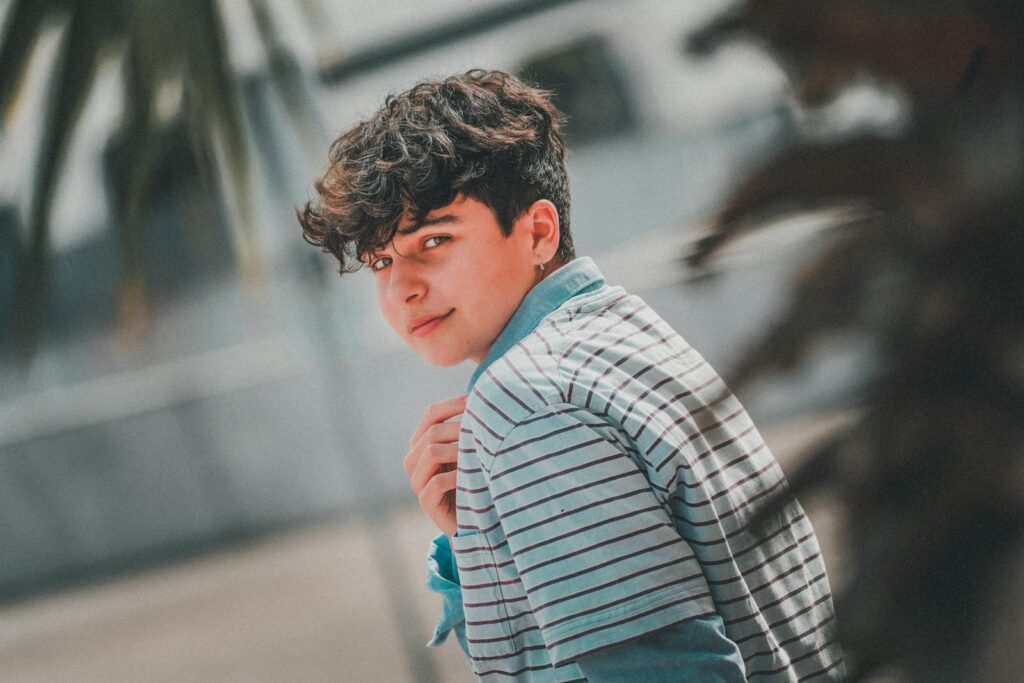 Side view of handsome male teenager with curly dark hair in stylish outfit chilling on street on sunny day and looking at camera