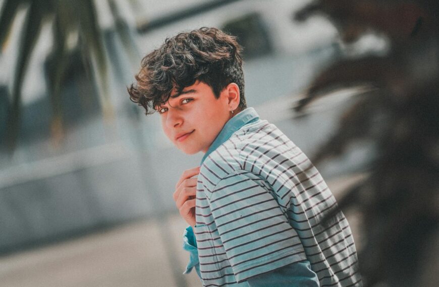 Side view of handsome male teenager with curly dark hair in stylish outfit chilling on street on sunny day and looking at camera